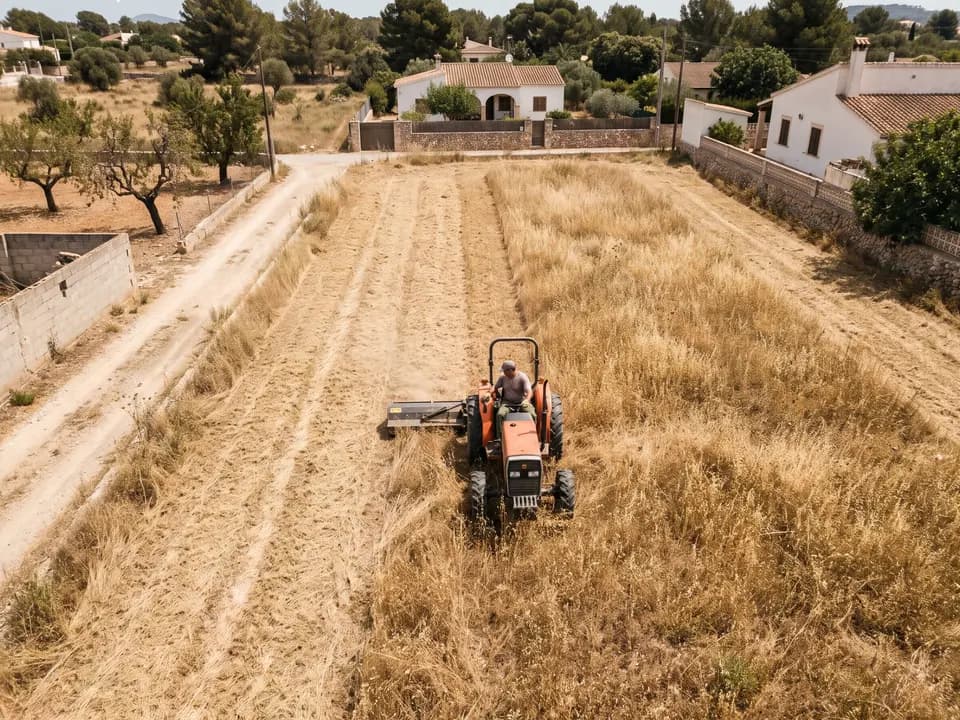 Tractor con desbrozadora de martillos cortando una parcela llana de hierba seca, visto desde un dron